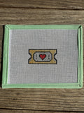 needlepoint design of a ticket with a red heart on a wooden surface