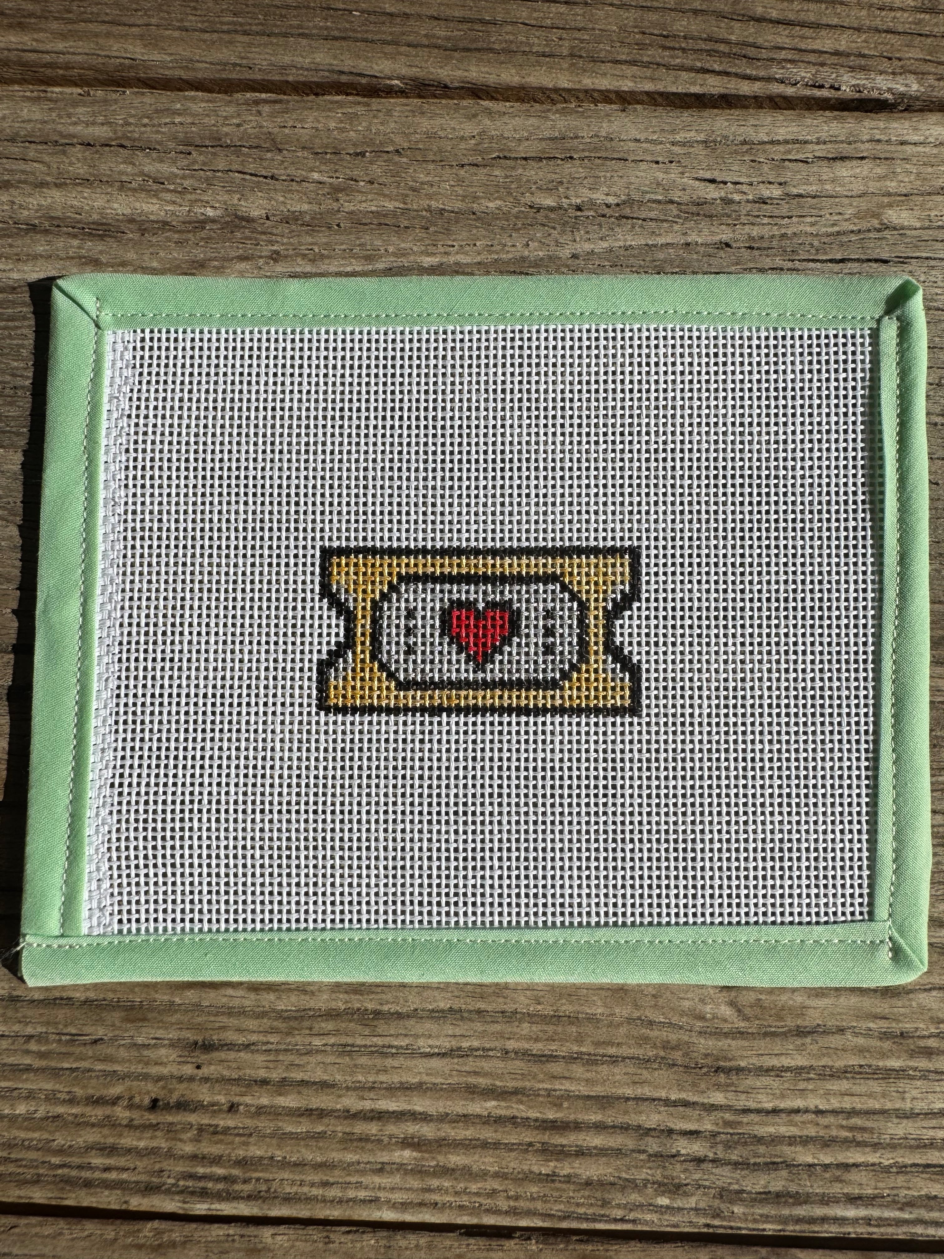 needlepoint design of a ticket with a red heart on a wooden surface