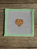 needlepoint heart  pizza slice on fabric with a green border on a wooden surface

