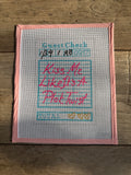 Cross-stitched piece on fabric with text and dates, framed in pink, on a wooden surface.
