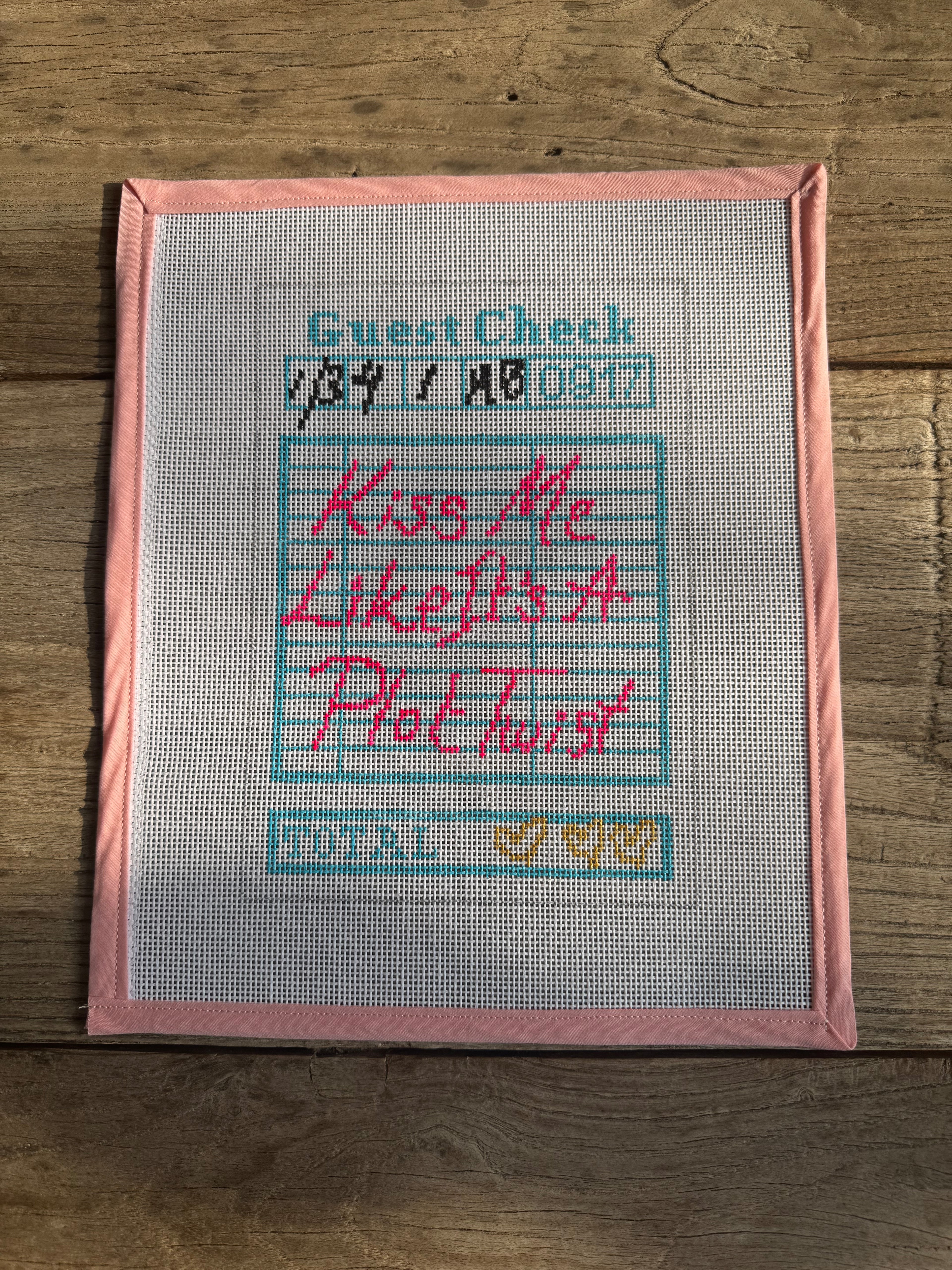 Cross-stitched piece on fabric with text and dates, framed in pink, on a wooden surface.