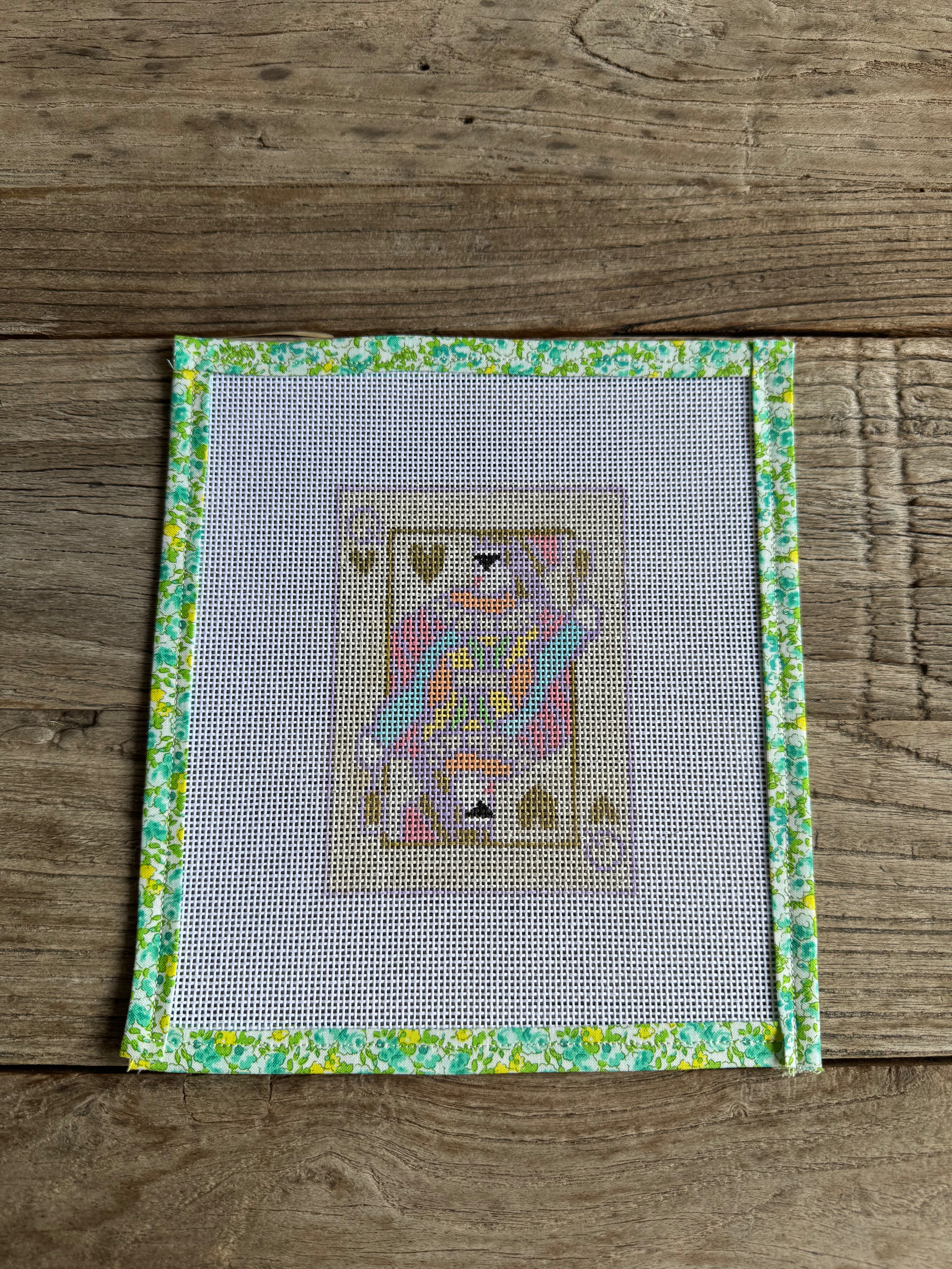Needlepoint design on fabric with a colorful queen of hearts pattern, framed by floral border, on wooden surface.
