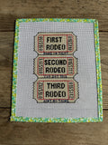 Cross-stitched pattern of rodeo tickets on fabric with a floral border.

