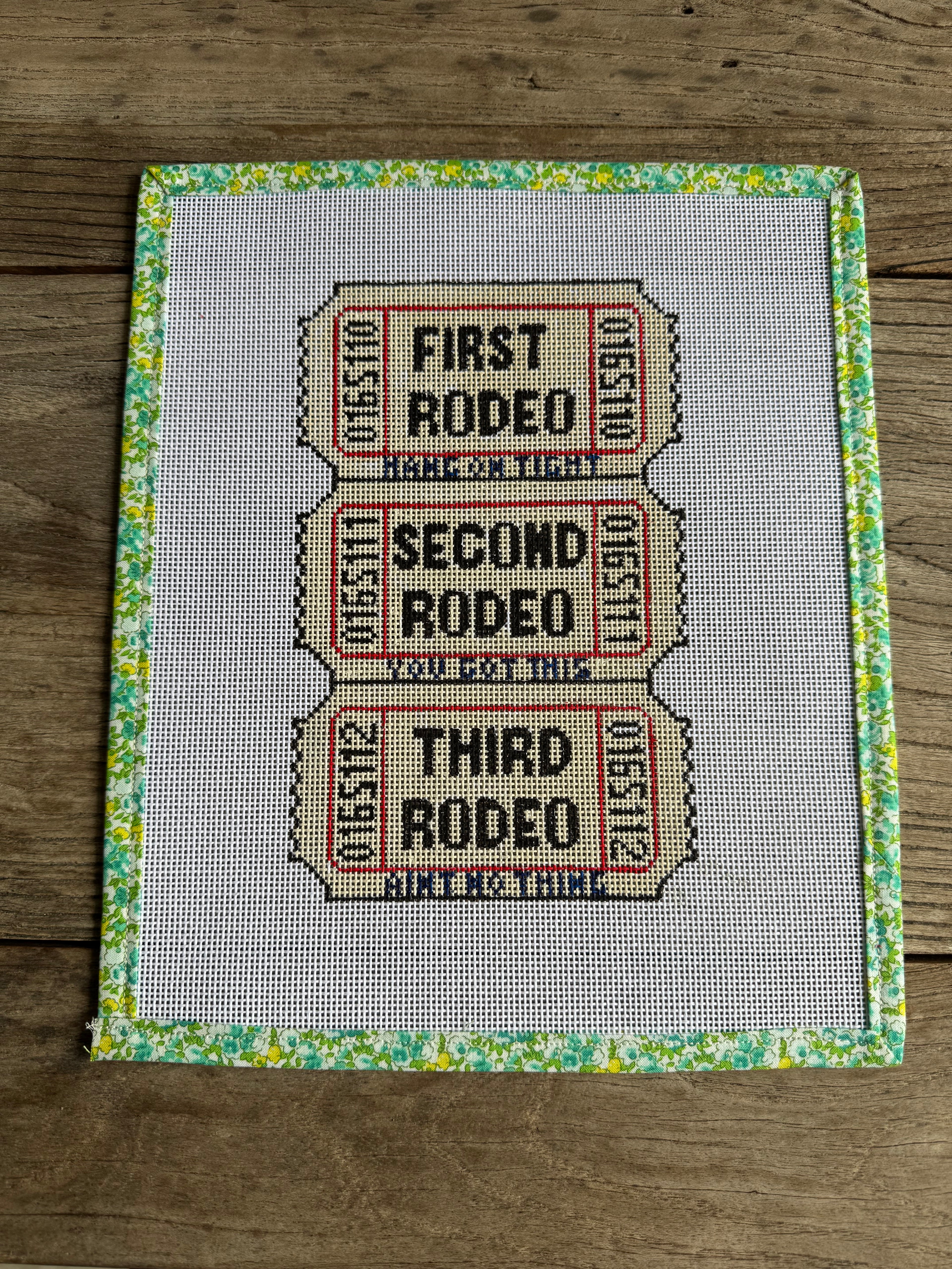 Cross-stitched pattern of rodeo tickets on fabric with a floral border.
