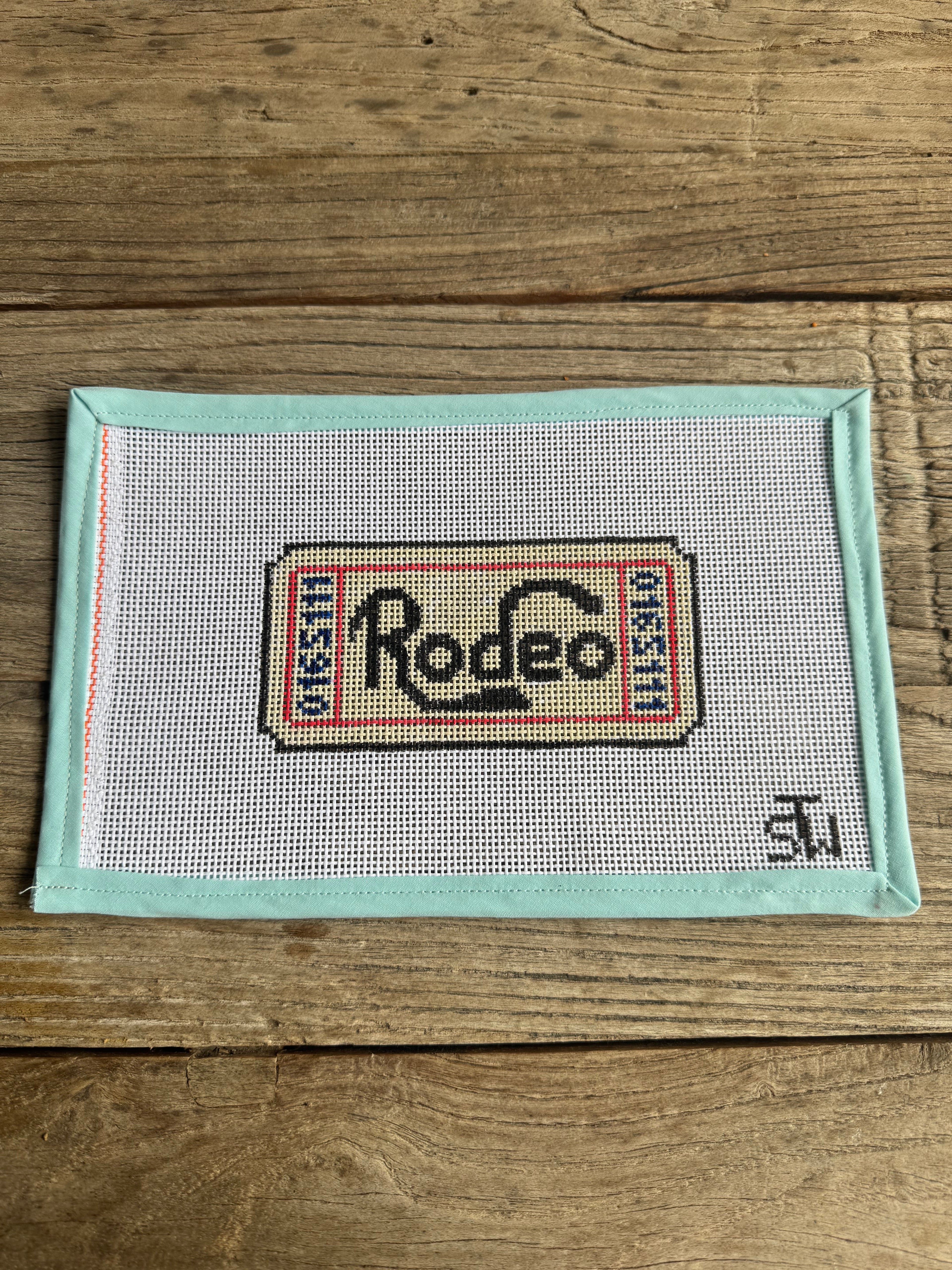 Cross-stitched design of a vintage Rodeo label on fabric with a light blue border.