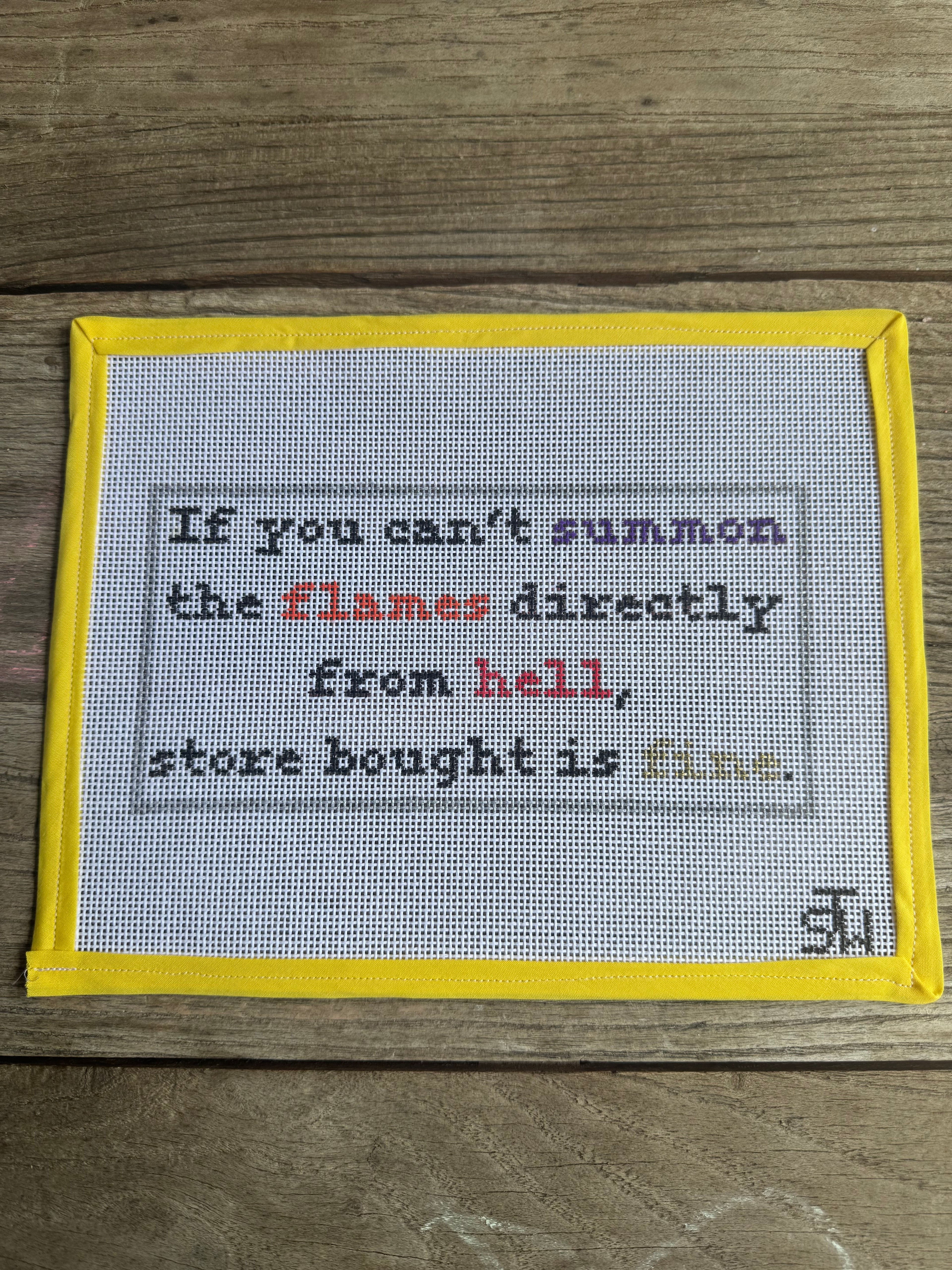 A square cross-stitch sampler with a yellow border, featuring a humorous embroidered quote on white Aida cloth, displayed flat on a rustic wooden background.