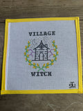 A square cross-stitch piece featuring a black house illustration surrounded by yellow and green floral details, with the words 'VILLAGE WITCH' stitched above and below. The fabric has a yellow border and is laid flat on a brown wooden surface.