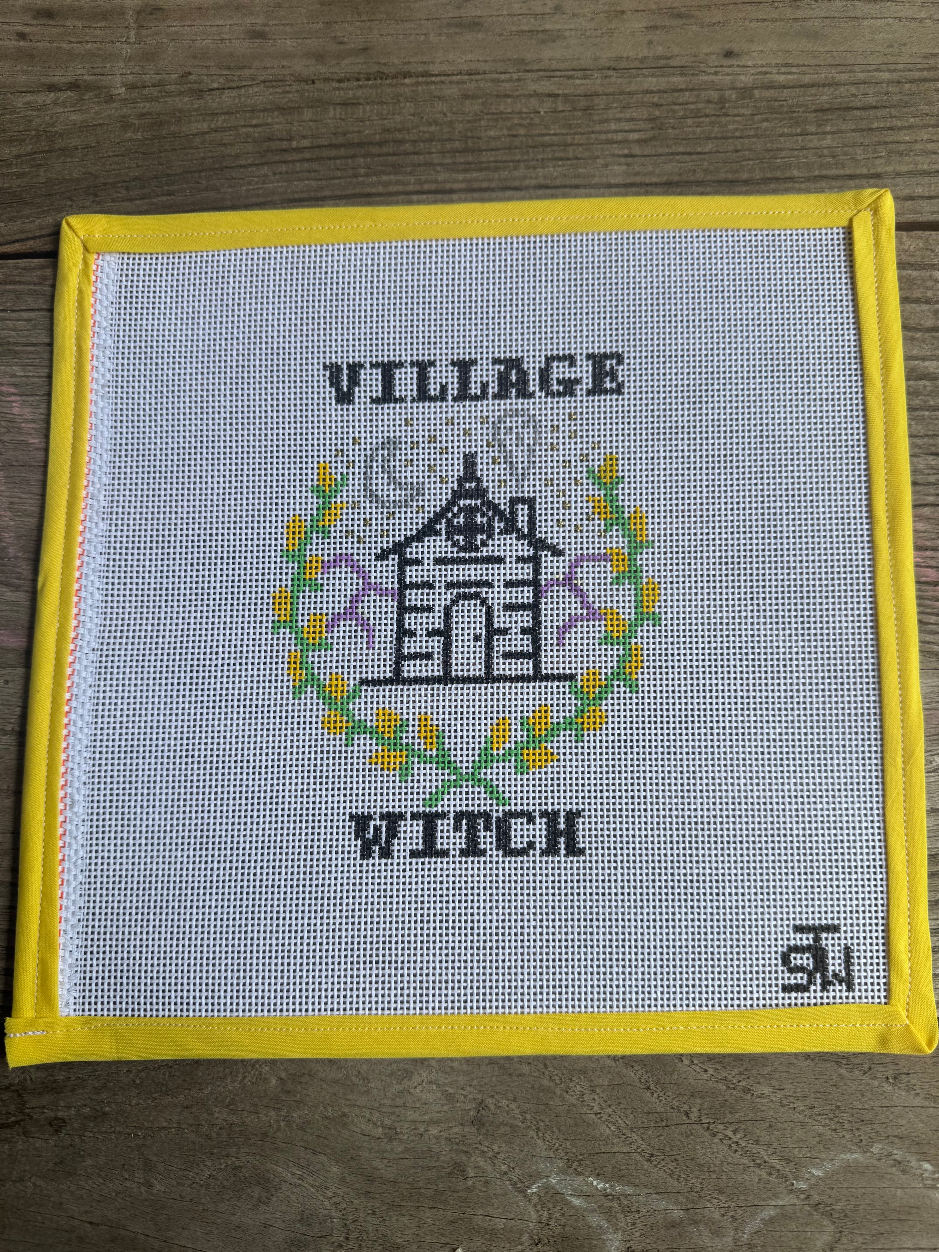 A square cross-stitch piece featuring a black house illustration surrounded by yellow and green floral details, with the words 'VILLAGE WITCH' stitched above and below. The fabric has a yellow border and is laid flat on a brown wooden surface.
